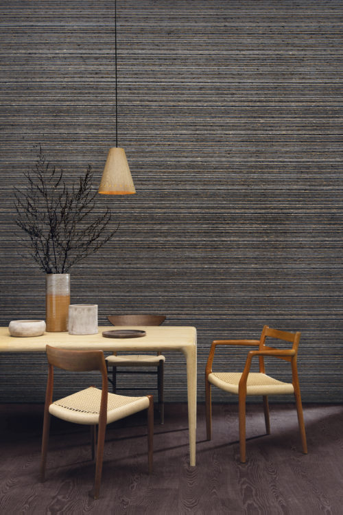 The Atelier collection by Omexco is an eclectic blend of artisanal craftsmanship and contemporary couture. On the wall of this inviting dining room we see a horizontally shown chunky weave in dark grey, brown and beige hues. The space is furnished with a Scandinavian style dining table and chairs, marked by a focus on clean, simple lines and minimalism. We also see a a chestnut parquet floor, a hanging lamp with wooden lightshade and ceramic vases and plates on the table.