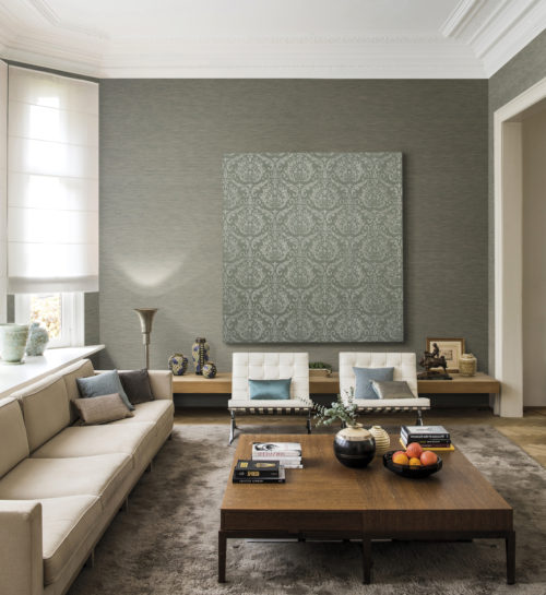 Living room with a high ceiling and big windows. 4 seater sofa and lounge chairs in cream. On the wall one plain wallcovering from the Elegance range and one damask installed on a frame. Standing out like a piece of art.