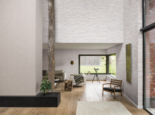 Living room with a lot of wood structures. There's a wooden floor but also a antique washed wooden beam in the middle of the room. A large window to look at the wildlife around. Shades of Pale wallcoverings complete the natural look.