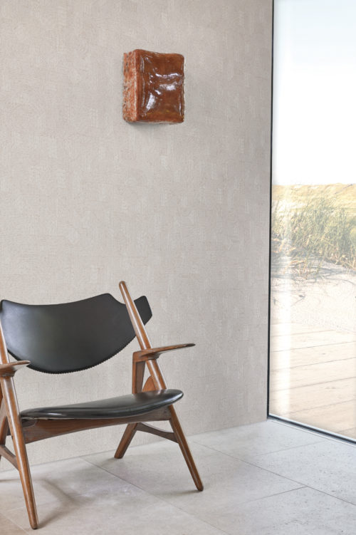A captivating view of Omexco's high-performance wallcovering collection, Rivage, adorning the wall behind a wooden armchair. The textured and sculpted motif in soothing beige tones evokes the intricacies of carved woodwork, creating an elegant backdrop. Through the window, dunes stretch out, adding a natural and serene touch to the scene. These PVC-free Rivage wallcoverings not only provide bleach cleanability but also feature enhanced scrubbability, seamlessly blending style and durability in this inviting and well-appointed space.