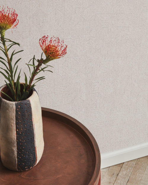 A close-up image featuring a brown round side table adorned with a ceramic vase holding two vibrant orange flowers. In the background, Omexco's high-performance wallcovering collection, Rivage, unfolds with a textured and sculpted motif reminiscent of intricately carved woodwork in soothing beige tones. These PVC-free Rivage wallcoverings not only offer bleach cleanability but also boast enhanced scrubbability, combining style and durability seamlessly in this elegant and artfully designed space.