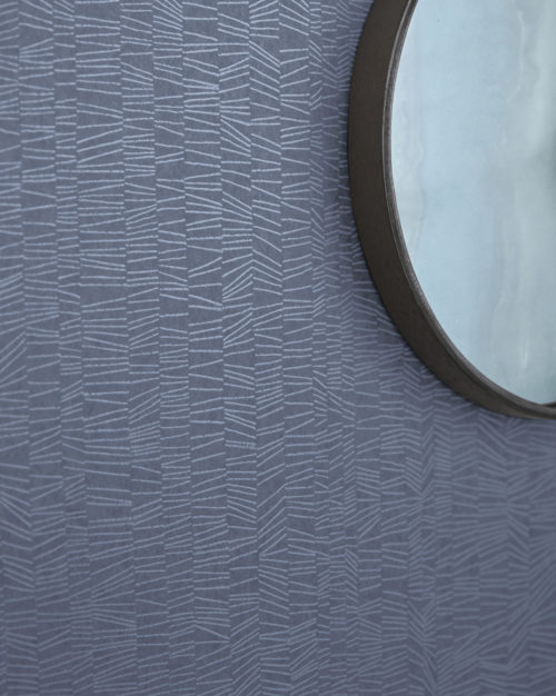 A captivating close-up of a wall adorned with Omexco's high-performance wallcovering collection Rivage in a soothing blue hue. The design boasts textured tonal stripes that beckon with subtle elegance. A round mirror hangs gracefully on the wall, reflecting the sophisticated ambiance. These PVC-free wallcoverings not only offer bleach cleanability but also feature enhanced performance with scrubbability, ensuring both style and durability.