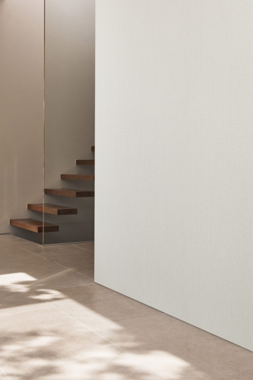 A close-up on a wall featuring Omexco's high-performance wallcovering collection, Rivage, showcasing a subtle plain white design. In the background, a floating wooden staircase adds a touch of modern elegance to this minimalist space. Large grey tiles cover the floor, and the interplay of shadows, cast by tree leaves in the beautiful daylight, adds a dynamic and serene ambiance. The simplicity of the design, coupled with the high-performance features of the Rivage wallcoverings, creates a visually striking and sophisticated atmosphere.