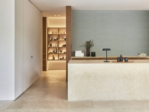 A tranquil and welcoming zen reception desk in soothing shades of beige, featuring a marble counter. In the background, a hanging open shelf cupboard adorned with decorative elements adds a touch of sophistication. The stone floor complements the serene atmosphere. A sleek black table lamp graces the marble counter. Behind the desk, Omexco's High Performance collection Rivage serves as a captivating backdrop, showcasing a light green design with a textured and sculpted motif, reminiscent of intricately carved woodwork, enhancing the overall sense of calm and elegance.