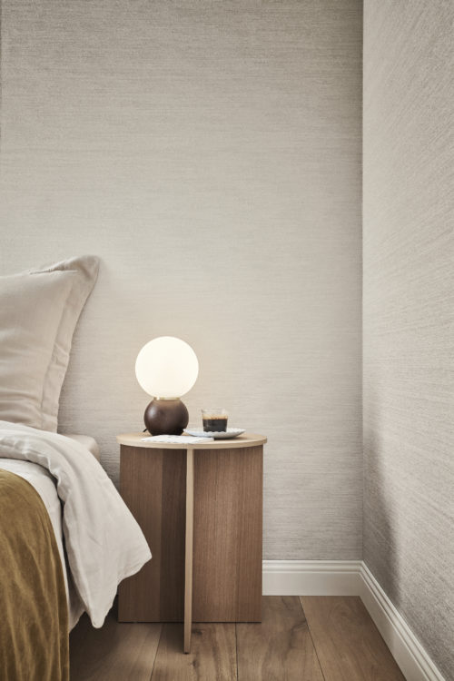 A cozy bedroom with a dark brown parquet floor, featuring a comfortable bed adorned with beige and ochre bedsheets. A wooden round bedside table holds a round table lamp. In the background, Omexco's textile wallcovering collection Café Society in light grey is displayed. Crafted with intricately structured and soft chenille yarn, this collection represents a modern revival of a textile classic.