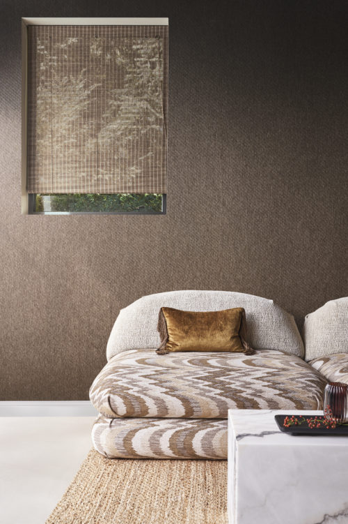 Here is a living room shot featuring a white cast floor and a natural jute braided carpet. Two comfortable couches with a brown print and a soft brown pillow complement the space. In the foreground, a marble coffee table stands out. The wall is adorned with embossed textile wallcoverings in chocolate brown from Omexco's Café Society collection, adding a timeless classic with a glossy flair that infuses the room with a luminous glow