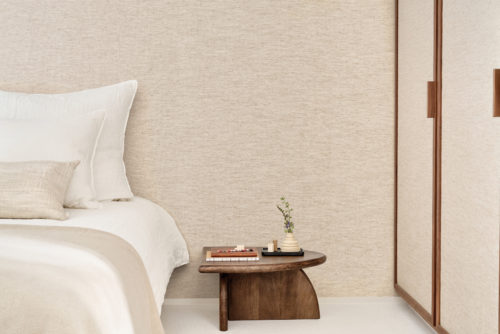 A serene bedroom interior featuring Omexco's Pure collection, reference PU333, a wallpaper that creates a subtle contrast between white yarn and natural-hued linen. The refined texture of the wallcovering adds warmth and depth, perfectly complementing the minimalist aesthetic. The scene includes a plush bed dressed in soft white and beige linens, paired with a matching textured pillow. Beside the bed, a sculptural wooden side table is adorned with stacked books, a small tray holding artisanal ceramics, and a delicate sprig of greenery in a vase. To the right, a wardrobe with wooden accents integrates seamlessly into the tranquil, natural-toned space, evoking a sense of comfort and understated elegance.