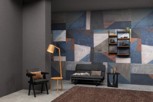 In this picture we see a feature wall with an XXL abstract art printed on fine mica. The geometric design with diagonal lines is pretty colorful with its shades of light and dark blue, beige, red and tones of grey. This is one of the designs of the Moonstone collection by Omexco, a Belgian brand of fine wallcoverings. In front of the wall there is a grey two seater sofa as well as a black chair with an orange pillow.