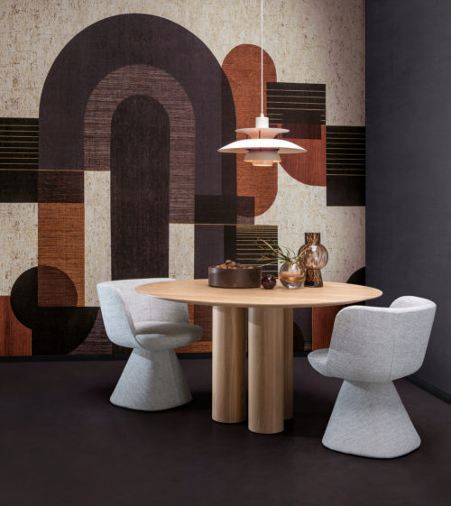 Example of a dining room with a bold graphic wallcovering showing a pattern of geometric shapes in grey, black, burnt orange and white hues. This panoramic design is digitally printed on cork and belongs to Omexco’s wallcovering collection “Casca”. We see a round wooden dining table in the center of the room with a white pendant lamp hanging above it. Decorative elements such as small glass vases and a bowl filled with walnuts are placed on the tabletop. Two soft grey dining chairs are positioned around the table. The overall aesthetic is modern with a touch of warmth and texture.