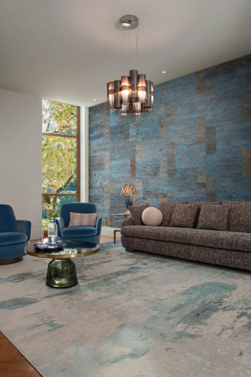 An image of a living room with bold, textured wallcoverings in shades of blue, which draws the eye towards the focal point of the room. These natural wallcoverings by Omexco have a leather-like look, but are actually made of Mulberry bark. The seating consists of a comfortable grey sofa and two stylish blue armchairs. The room is well-lit with natural light from a large window and a ceiling light fixture. Decorative elements such as a large rug in blue tones, a round coffee table in green glass and an organic shaped table lamp add interest and balance to the space.