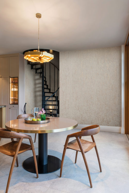 Example of a bright and airy kitchen featuring large light grey tiles, white kitchen cabinets, a round dining table with three wooden chairs and a black spiral staircase in the background. A gold pendant lamp adds a touch of elegance and warmth to the space, while a beige metallic wallcovering adds subtle texture and visual interest. Here we see Omexco’s cork wallcovering collection “Casca”. The overall effect is a modern and inviting kitchen that is perfect for both meal prep and casual dining.