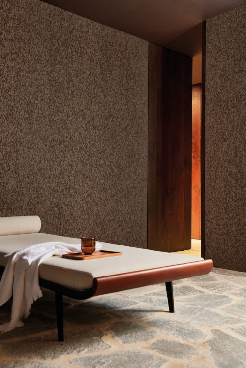 An image of a serene, neutral-toned room featuring textured wallcoverings with a brown organic pattern, a white daybed with a soft blanket. These natural wallcoverings have a leather-like look, but are actually made of cork oak bark. They belong to the exquisite, handmade “Casca” collection by Omexco. The textured walls add warmth and depth to the room, while the white daybed provides a comfortable seating option. The natural stone floor adds a touch of rustic charm and earthiness to the space. The overall effect is a calming and inviting environment perfect for relaxation or contemplation.