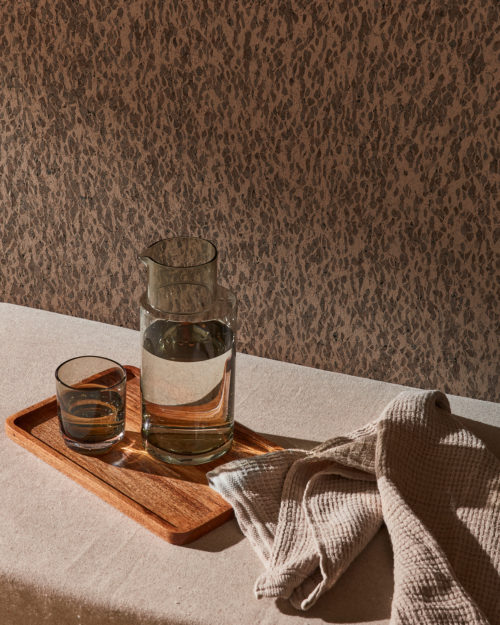Closeup of a small wooden tray with a transparent jug and a glass, both filled with water, and a beige kitchen towel. The items are placed on a soft daybed, also in beige. On the wall of this serene, neutral-toned room, we see a textured wallcovering with a brown organic pattern. These natural wallcoverings have a leather-like look, but are actually made of cork oak bark. They belong to the exquisite, handmade “Casca” collection by Omexco. The interesting play of natural daylight and shadow create a calming and inviting atmosphere.