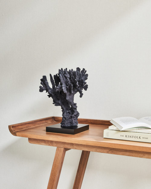 A close-up view of a wall adorned with Omexco's high-performance wallcovering collection, Rivage, showcasing a subtle plain white design from the collection. Against the wall, a Scandinavian-style wooden side table holds three books as well as a captivating coral sculpture. The PVC-free Rivage wallcoverings not only offer bleach cleanability but also feature enhanced performance with scrubbability, ensuring both style and durability.