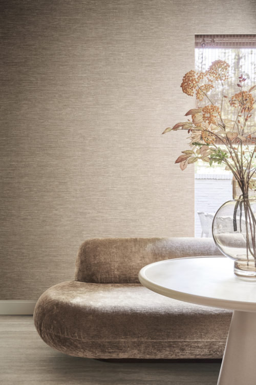 A living room shot featuring a white round table in the front with a transparent glass vase holding flowers on top of it. Behind the table is a very comfortable soft couch in light brown velvet fabric. As a backdrop, we see the textile wallcovering collection Café Society in shades of soft pink. This collection is a reinterpretation of one of Omexco's most iconic classics, embodying the luxurious essence of silk through meticulously printed textile fibers. The window on the right offers a view of an outdoor seating area.