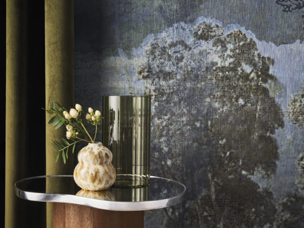 A decor shot featuring an olive-colored velvet curtain on the left. Next to it, an organically shaped side table holds a green vase and a smaller ceramic vase. The backdrop showcases a night landscape with trees from Omexco's Café Society wallcovering collection in shades of blue. Delicately printed on fine linen, this panoramic design evokes the grandeur of the past while embracing the essence of contemporary design.