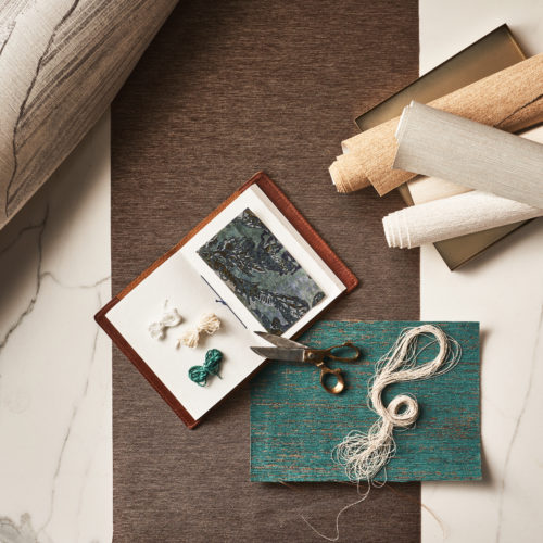 Material moodboard featuring brown, beige, and turquoise samples from Omexco’s textile wallcovering collection, Café Society. The samples, along with an open notebook, textile yarns, and scissors, are displayed on a marble background. The Café Society range distills the essence of a glamorous bygone era in its exclusive designs, featuring rich, velvety weaves, luxurious chenille yarns, and printed textiles.