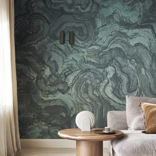 A living room decorated with a modern aesthetic. On the left, a large window framed by curtains allows natural light to flood the space. To the right, a comfortable grey couch is adorned with a brown pillow, creating a cozy atmosphere. Beside the couch is a round wooden side table, adding a touch of rustic charm. The floor is made of light wood, complemented by a brown carpet that grounds the room. The backdrop features Omexco's teal printed textile yarns from the Café Society collection, evoking the subtle, intricate patterns of malachite, reminiscent of nature’s captivating artistry.