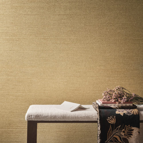 A home decor closeup featuring a dark wooden seat bench with a soft white cushion positioned on a grey cast floor. On top of the seat bench are some books, fresh flowers, and a soft blanket with a floral print. In the background, Omexco's textile wallcovering collection Café Society in golden brown is displayed. Crafted with intricately structured chenille yarn, this is a modern revival of a textile classic.