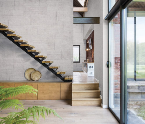 Wall with raffia blocks wallcovering. Stairs run diagonally through the picture. Large window on the right. We get a look at the tv room in the back with brick wall and bird statues.