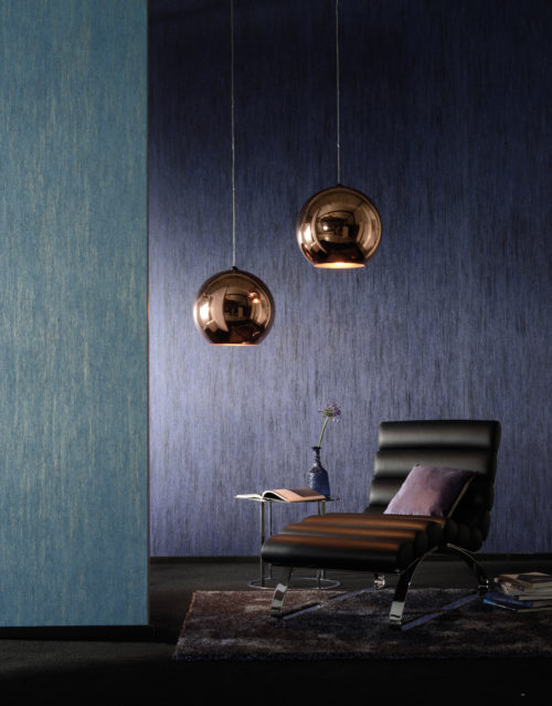 Well balanced room with a fluffy carpet, leather chaise longue and two wallcoverings. One purple, one petrol green. Two copper lamps reflect the little light there is. On a table next to the chaise a vase with a purple flower.