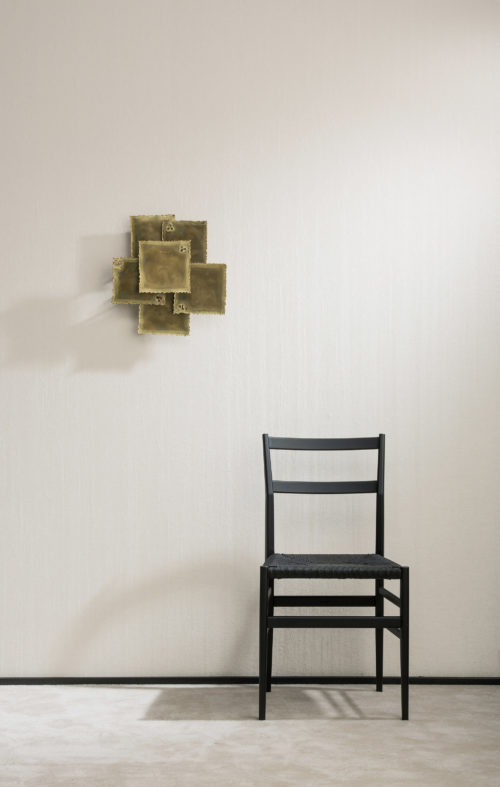 A lone black chair stands next to an art piece that unfolds on the wall. Crushed linnen Omexco wallcovering.