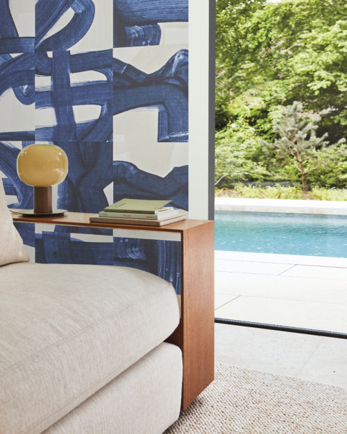 A stunning living room featuring Omexco's Rivage wallcovering in a captivating blue hue with artistic brushstrokes. The design skillfully weaves echoes of ropes and maritime knots, paying tribute to the intricacies of seafaring. In the foreground, a close-up of a beige sofa and a minimalist wooden side table exudes modern elegance. Beyond, an open window reveals a refreshing view of a swimming pool, adding to the overall sense of tranquility and sophistication in this beautifully adorned space.