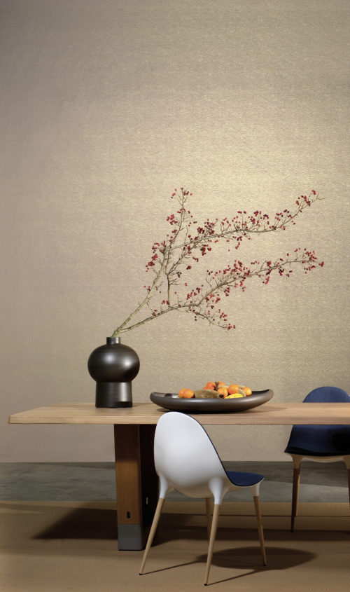Light brown and gold wallcovering set the soft tone in this dining room. Chairs are white plastic with bleu fabric. The table is dressed with a huge fruit bowl. Vase with berry branches.