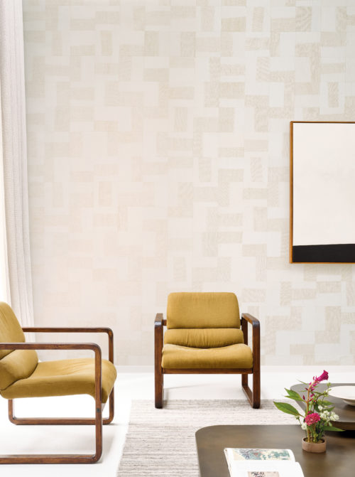 A modern and inviting living room showcasing Omexco's Pure collection, reference PU261, named Intensify. This geometric paper patchwork wallpaper is enriched with linen-toned openwork abaca weaving, creating a design that exudes depth and texture. The wallpaper’s subtle yet intricate patterns harmonize beautifully with the room’s minimalist aesthetic. Two mid-century modern armchairs upholstered in mustard-yellow fabric bring warmth and contrast to the neutral tones of the wall. A sleek coffee table adorned with a vibrant floral arrangement and books adds personality to the space. The abstract framed artwork on the wall complements the structured yet organic design of the wallpaper, making the room feel contemporary and stylish.