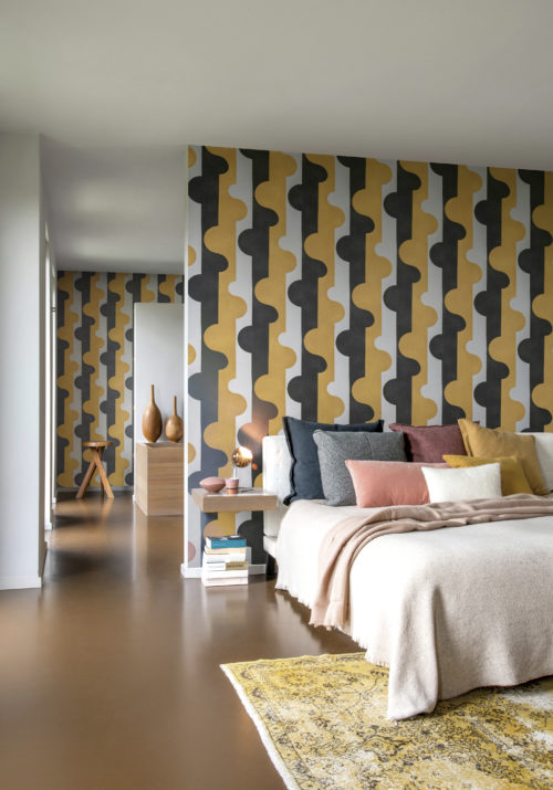 Modern, inviting bedroom with a yellow carpet, colourful pillows on the bed and floating bedside tables. On the walls of this contemporary space, we see the Puzzle of Passion design from Omexco's wallcoverings collection Joy. The playful design shows a puzzle-like pattern in contrasting shades of yellow, black and white.