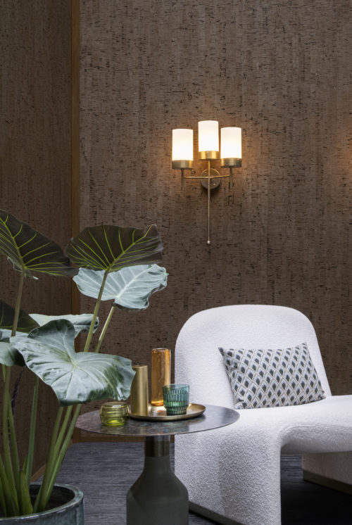 An image of a cozy living room corner featuring dark brown wallcoverings, a tropical houseplant in the foreground and a white soft single seater with a decorative pillow on the right. A round side table provides a surface for small glass vases while a 3-lights wall lamp in gold adds a touch of sophistication and elegance to the space. The wallcoverings belong to the exquisite collection “Casca” by Omexco, exposing the soft and matt natural textures of cork. This cork design is reminiscent of the look of birch bark.
