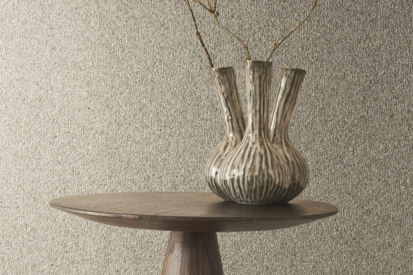 A charming decor composition is captured in this photo, featuring a wooden side table adorned with a distinctive fennel-shaped vase. Dried flowers grace the vase, adding a rustic touch. The backdrop showcases the elegance of Omexco's Archipelago collection, featuring embossed grey mica wallcoverings that contribute to the overall sophistication of the scene, creating a harmonious blend of natural elements and contemporary design.