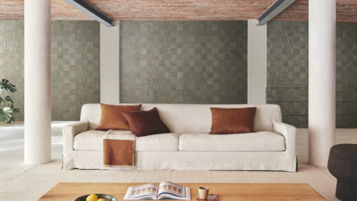 This image showcases a spacious loft-like living room with a light grey cast floor. Two stone columns frame the room, adding an architectural element. In the background stands a vibrant Monstera plant, adding a touch of greenery to the space.  In the foreground, a large beige sofa adorned with brown pillows takes center stage, positioned on a beige carpet. A wooden coffee table sits in front of the sofa, displaying a book, a cup, and some lemons.  The focal point of the room is Omexco's handmade natural wallcoverings collection "Intuition". The wall is adorned with a textured cattail patchwork in green, featuring a chequered pattern.