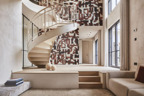A captivating living room in soothing shades of beige, featuring a central spiral staircase and large windows on the right. Behind the staircase, an eye-catching wall adorned with Omexco's Rivage collection, a non-woven wallcovering showcasing a stunning print of brown brushstrokes on a white background. The design skillfully evokes the textures of ropes and maritime knots, paying homage to the intricacies of seafaring.