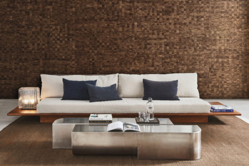 Stylish living room boasting a gray cast floor and a plush brown carpet. The centerpiece is a large minimalist sofa with a wooden frame, adorned with light beige cushions and dark blue pillows. Flanking the sofa are glass table lamps and piles of books on each side. In front of the sofa, a silver-colored metal coffee table displays more books. The true focal point is the striking wall adorned with Omexco's handmade natural "Intuition" wallcoverings. The mosaic-like mulberry weave in shades of brown adds a captivating and textured backdrop, enhancing the room's sophistication.