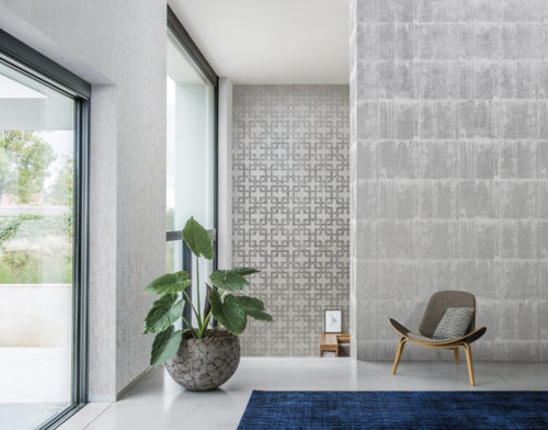 Well lit room with 3 walls of cork wallcovering. One with silver blocks, one with a silver geometrical design and one plain with golden dots. Armchair and large plant, bleu carpet.
