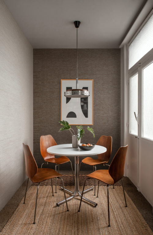 In this image we see a cosy dining nook with a vintage round dining table and retro wooden chairs, a pendulum lamp and ethnic woven carpet. On the walls, we see beige and brown paper yarns delicately woven into seamless wallcoverings by Omexco. Design 'Flow' from the wallcovering collection Tribu.
