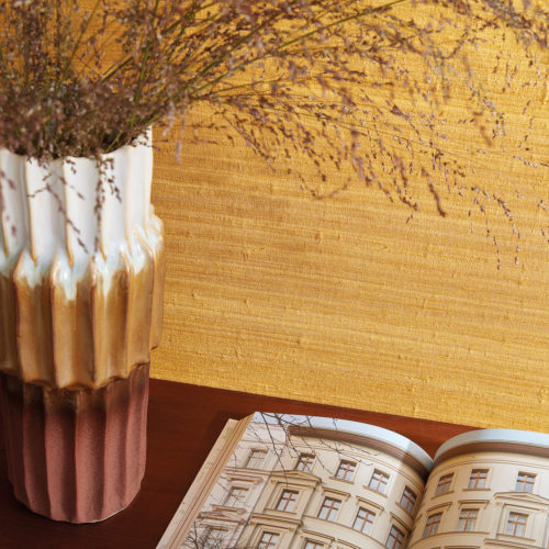Closeup of an open architecture book laying next to a tricolor ceramic 70's vase. The vase is holding some dried flowers. Both items are placed on a wooden table or sideboard, standing against an ochre wall with a precious silk textile design. These are Omexco’s "Loom Stories” wallcoverings, a rich and eclectic collection of irregular textures made from hand-spun yarns, fine Belgian linen and wild silk, all in a diverse palette that ranges from soothing neutrals to eye-catching, vivaciously bold colours.