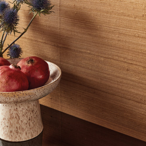 Closeup of a speckled ceramic 70's bowl on foot on which two pomegranates are displayed. Next to the bowl we see some blue dried flowers. The items are placed on a wooden table or sideboard, standing against an terracotta coloured wall with a precious silk textile design. These are Omexco’s "Loom Stories” wallcoverings, a rich and eclectic collection of irregular textures made from hand-spun yarns, fine Belgian linen and wild silk.