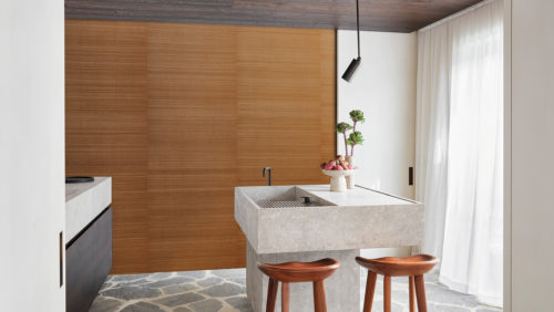 A bright and airy kitchen featuring a natural stone floor that adds a touch of earthy elegance. The walls are adorned with a stunning burnt orange silk wallcovering from Omexco’s textile collection ‘Loom Stories’. In the center of the room, there is a beautiful kitchen island made of stone, with a bowl of fresh vegetables and a vase with flowers on top. Two wooden barstools are placed around the island, providing comfortable seating for family and friends. The combination of natural materials, vibrant colors and functional design makes this kitchen a warm and inviting space for cooking and gathering.