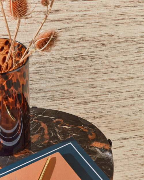 A closeup photo of a stylish marble side table with a speckled glass vase filled with dried flowers on top of it. An orange notebook is placed next to the vase, and its bright orange color pops against the neutral backdrop. Here we see Omexco's Loom Stories wallcoverings in shades of cream and light brown, with a subtle pattern adding texture to the wall. The wild aesthetic is created by the irregularity of the silky yarns.