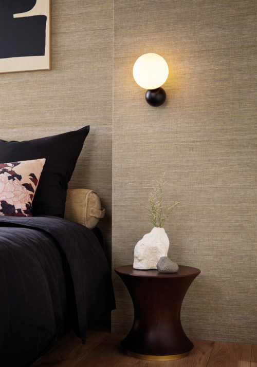 An image of an inviting bedroom showcasing a cozy bed with a pink floral pillow. Beside it, a wooden round side table with a minimalist bulb wall Lamp hanging above it provides warm ambient lighting. The wooden floor complements the natural textures of the room, while the wall features stunning straw-colored silk wallcoverings by Omexco, adding a touch of luxury and sophistication to the stylish decor. This textile wallcovering collection is called Loom Stories.