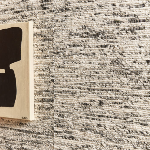 Closeup on a wall with Omexco’s "Loom Stories” collection, a range of thickly woven textile wallcoverings, shown here in off-white and black shades. On the image we see also a part of a modern, minimalistic black painting on white canvas.