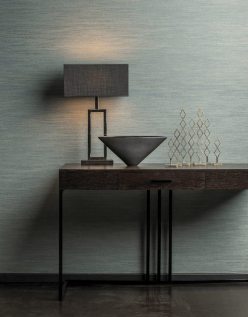 Green Omexco wallcovering, installed horizontally. In front of it a table lamp with grey shade on a desk with one drawer and black feet. Metal artwork with diamond shapes.