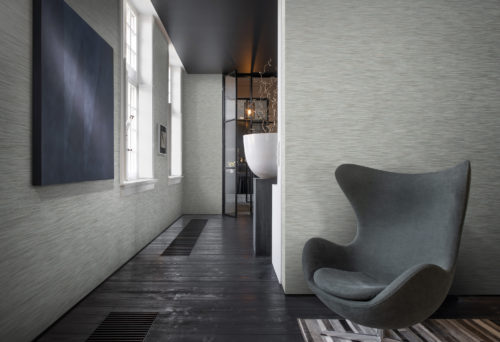Room with black wooden floor and black iron door in the back. In the front a grey armchair. Classic Omexco wallcoverings on the wall.