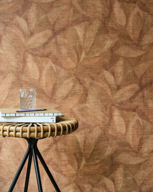 This inviting interior highlights Omexco’s OAS202 reference, Hierbas, from the Oase collection. The wallcovering features a delicate leaf motif, printed onto natural raffia, evoking an organic and warm atmosphere. The earthy terracotta tones of the wall harmonize with the rattan and metal side table, creating a setting that feels both rustic and refined. A stack of books, a glass of water, and a pen placed atop the table enhance the relaxed yet sophisticated ambiance, making this space ideal for moments of calm and contemplation.