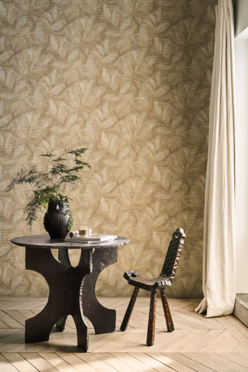 This serene interior highlights Omexco’s OAS201 reference, Hierbas, from the Oase collection. The wallcovering features an organic leaf pattern, delicately printed onto a natural raffia weave, evoking a sense of tranquility and harmony with nature. The warm, earthy tones of the wall are complemented by a sculptural black wooden chair and a textured round table, creating a rustic yet refined atmosphere. A decorative owl-shaped vase filled with lush greenery adds a touch of life, while the flowing linen curtain allows natural light to softly filter into the space, enhancing its serene and organic essence.