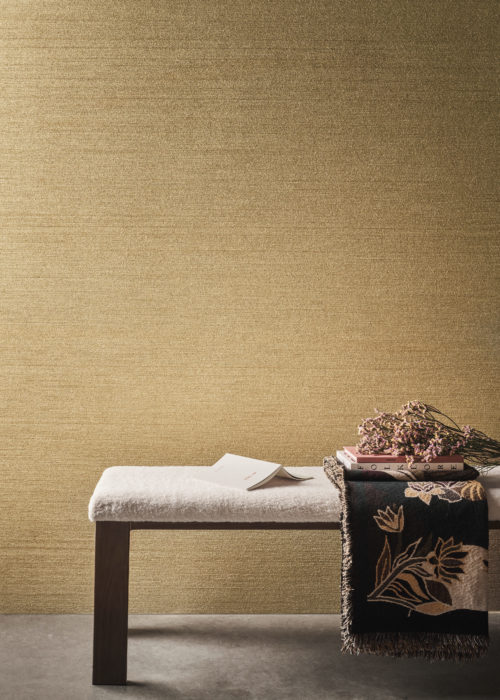 A home decor closeup featuring a dark wooden seat bench with a soft white cushion positioned on a grey cast floor. On top of the seat bench are some books, fresh flowers, and a soft blanket with a floral print. In the background, Omexco's textile wallcovering collection Café Society in golden brown is displayed. Crafted with intricately structured chenille yarn, this is a modern revival of a textile classic.