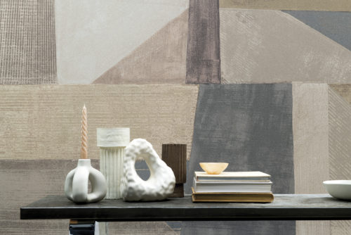 Moonstone by Omexco is a lavish collection of textured wallcoverings softly iridescent like the Moonstone. In this picture we see a feature wall with an XXL abstract art printed on fine mica. The geometric design with diagonal lines is colorful, yet not overwhelming with its soft shades of beige, white, grey, soft pink and unsaturated blue. In front of the wall, we see a low wooden coffee table with small vases and other ceramic decorations as well as a pile of books.