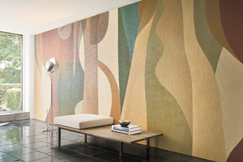 This stunning interior highlights the captivating OAS211 reference, titled Alegre, from Omexco's Oase collection. The wallcovering is crafted from printed paperweave, showcasing a sweeping panoramic design inspired by naïve art, evoking the vibrant landscapes of South America. The interplay of warm earth tones and organic shapes brings life and depth to the space. Positioned against this statement wallpaper is a minimalist bench with a natural wooden slat design, topped with a neutral cushion for added comfort. A sleek floor lamp with a metallic finish elegantly complements the setting, while the glossy black tiles on the floor add a modern and grounding element. The large window on the left invites natural light, seamlessly connecting the indoor atmosphere with the greenery outside, creating a harmonious and uplifting environment.